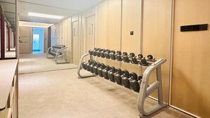 A hotel corridor with a row of dumbbells on a rack, a mirror reflecting the corridor, and a door at the end.