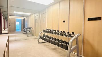 A hotel corridor with a row of dumbbells on a rack, a mirror reflecting the corridor, and a door at the end.