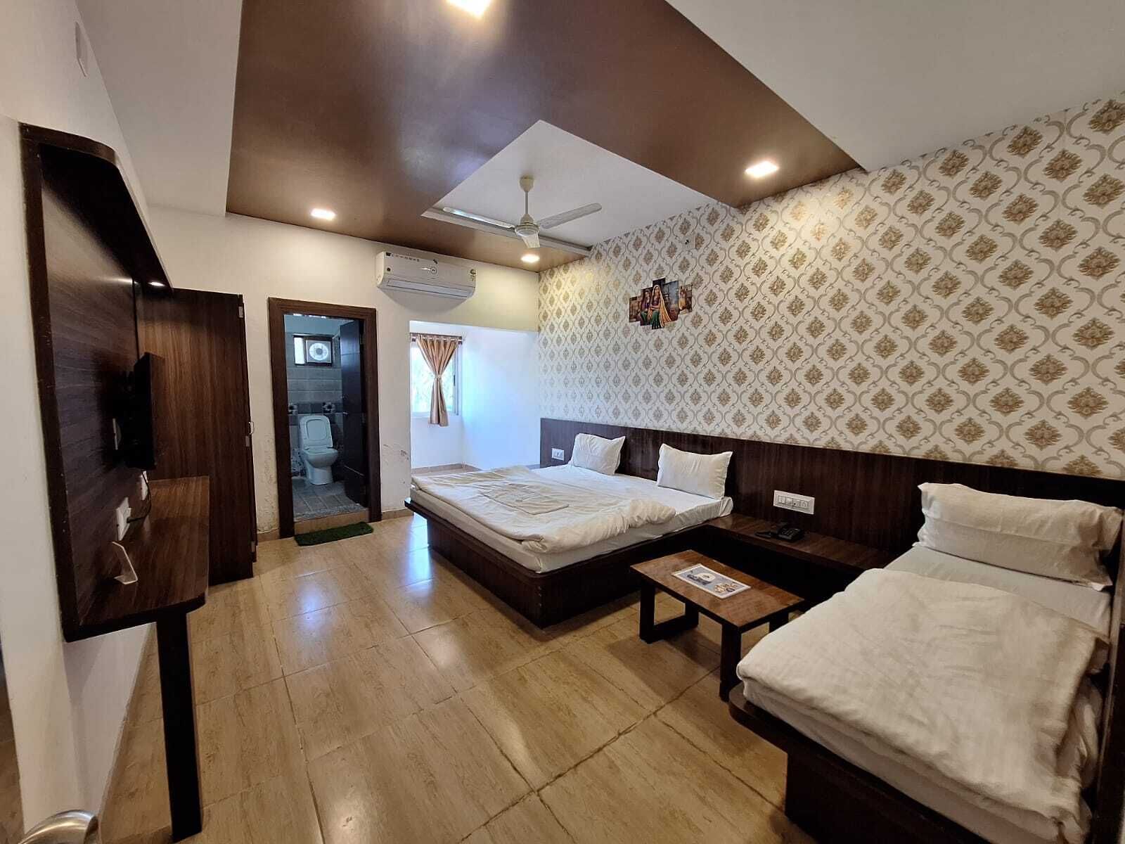 Quarto de hotel com uma cama grande, uma cama menor, uma escrivaninha, um guarda-roupa e um banheiro com vaso sanitário e chuveiro.