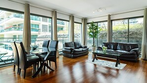 A spacious living room with a glass dining table, two leather sofas, and a coffee table. Large windows offer a view of a building and greenery outside. - GuestReady - Relaxing Funchal Retreat (Funchal)