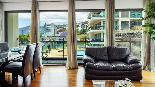 A modern living room with a leather sofa, a dining area with a glass table and chairs, large windows with curtains, and a view of a pool and buildings. - GuestReady - Relaxing Funchal Retreat (Funchal)