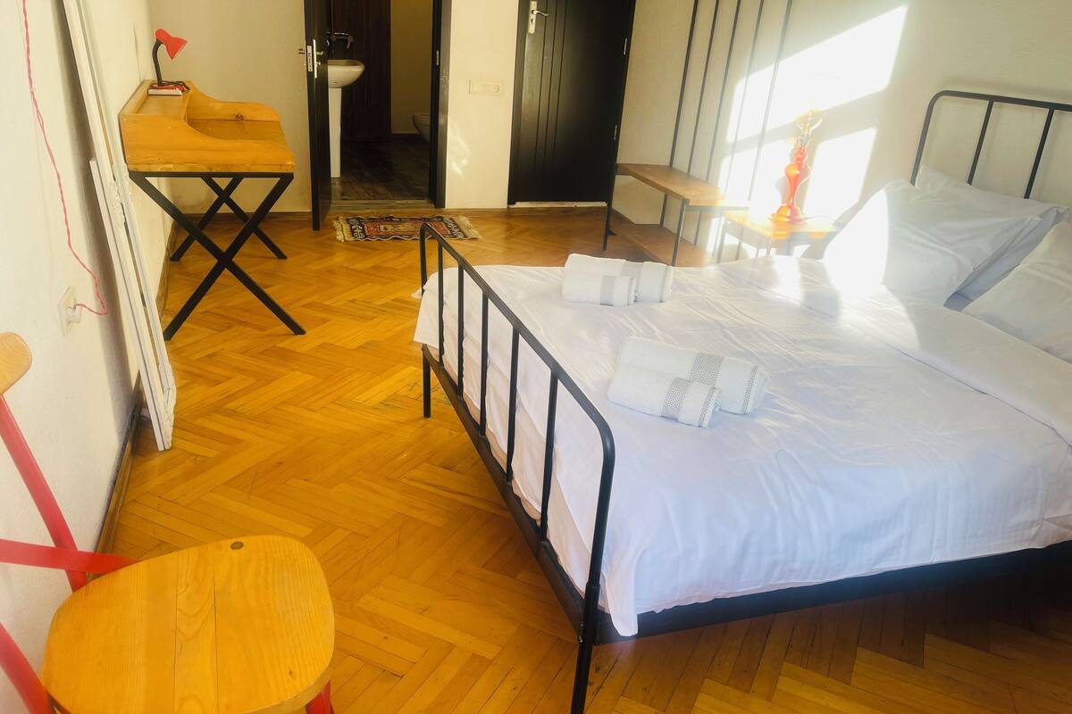 A bedroom with a wooden floor, a bed with white linens, a small wooden table, and a red chair.