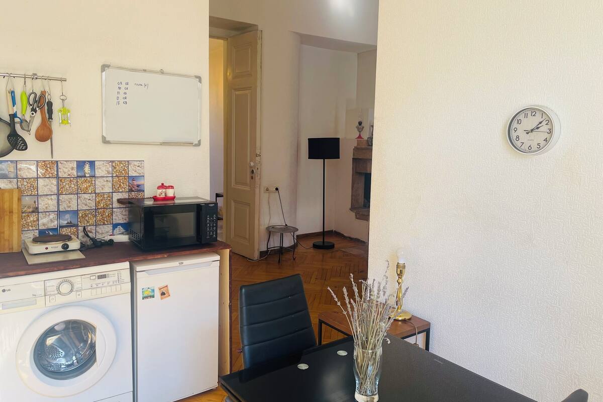 A compact kitchen with a microwave, washing machine, and a whiteboard. There's a dining table with a vase of dried flowers.