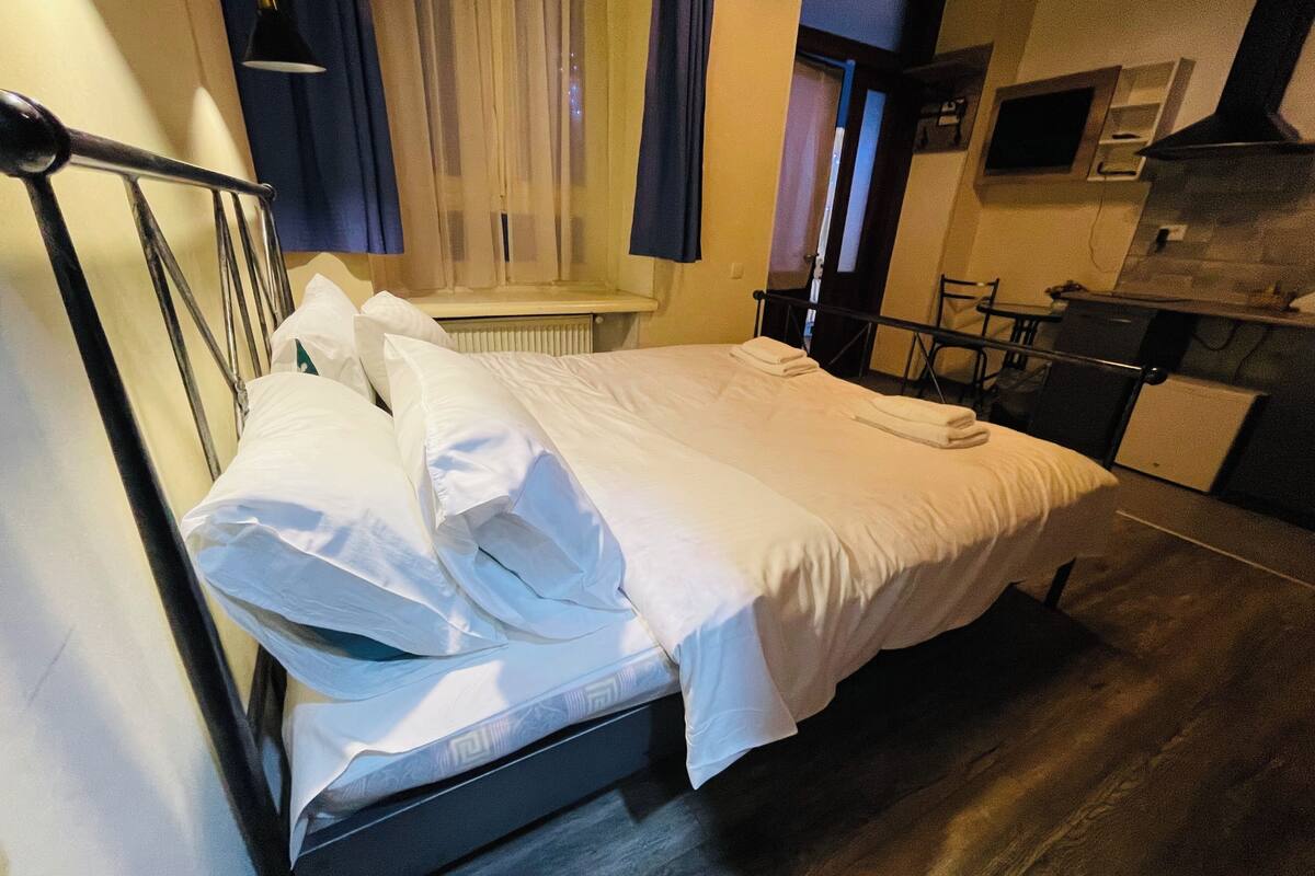 A neatly made bed in a hotel room with a wooden floor, a radiator, and a small kitchenette area.