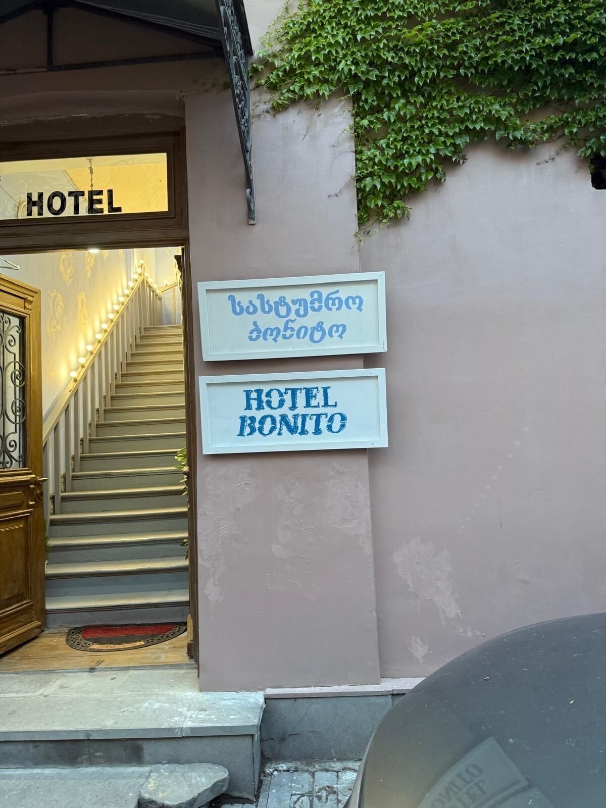 A hotel entrance with a staircase leading up to a lit interior. There are signs indicating the hotel name in English and another language.
