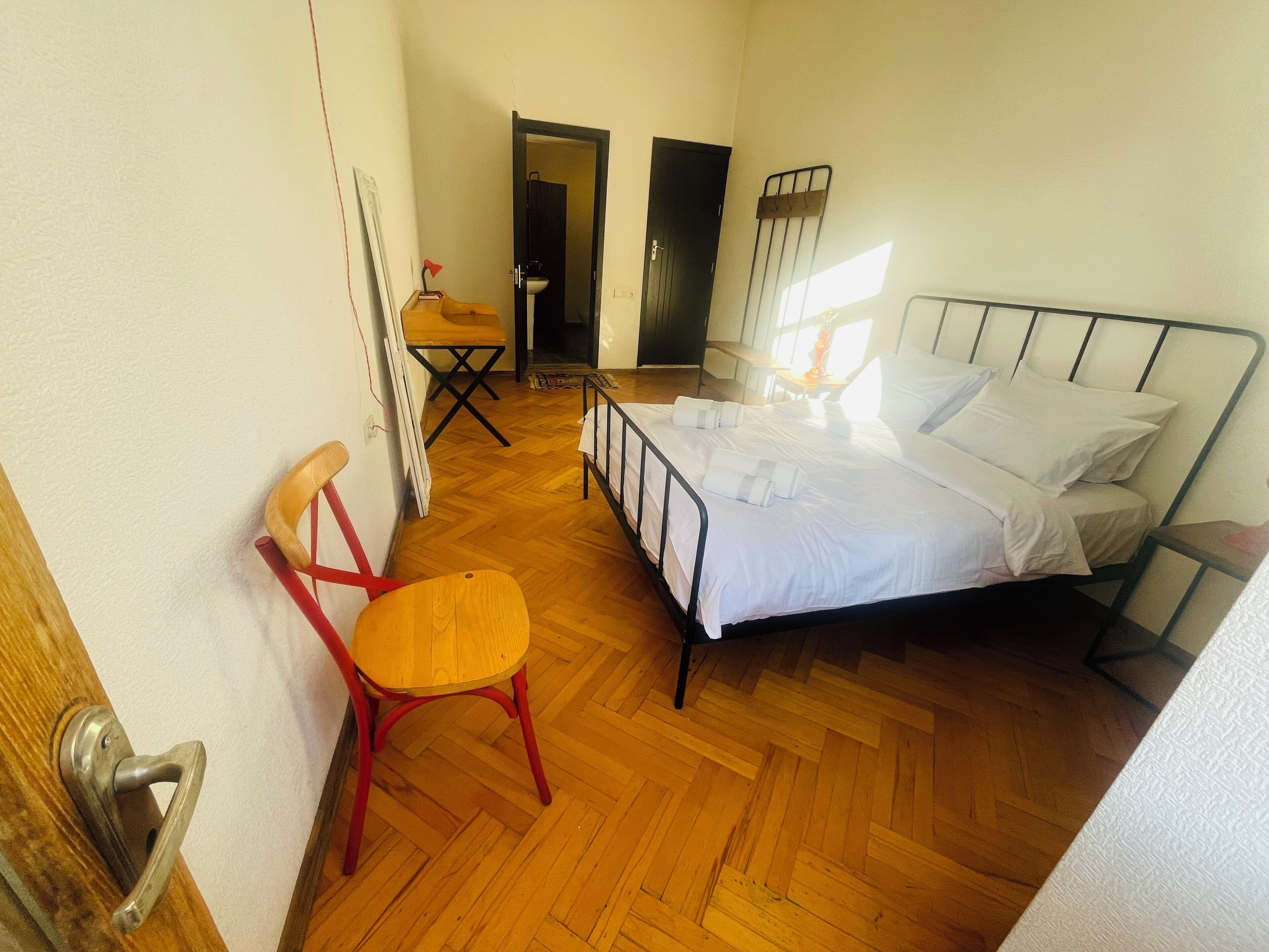 A bedroom with a bed, a chair, a small table, and a door leading to another room.