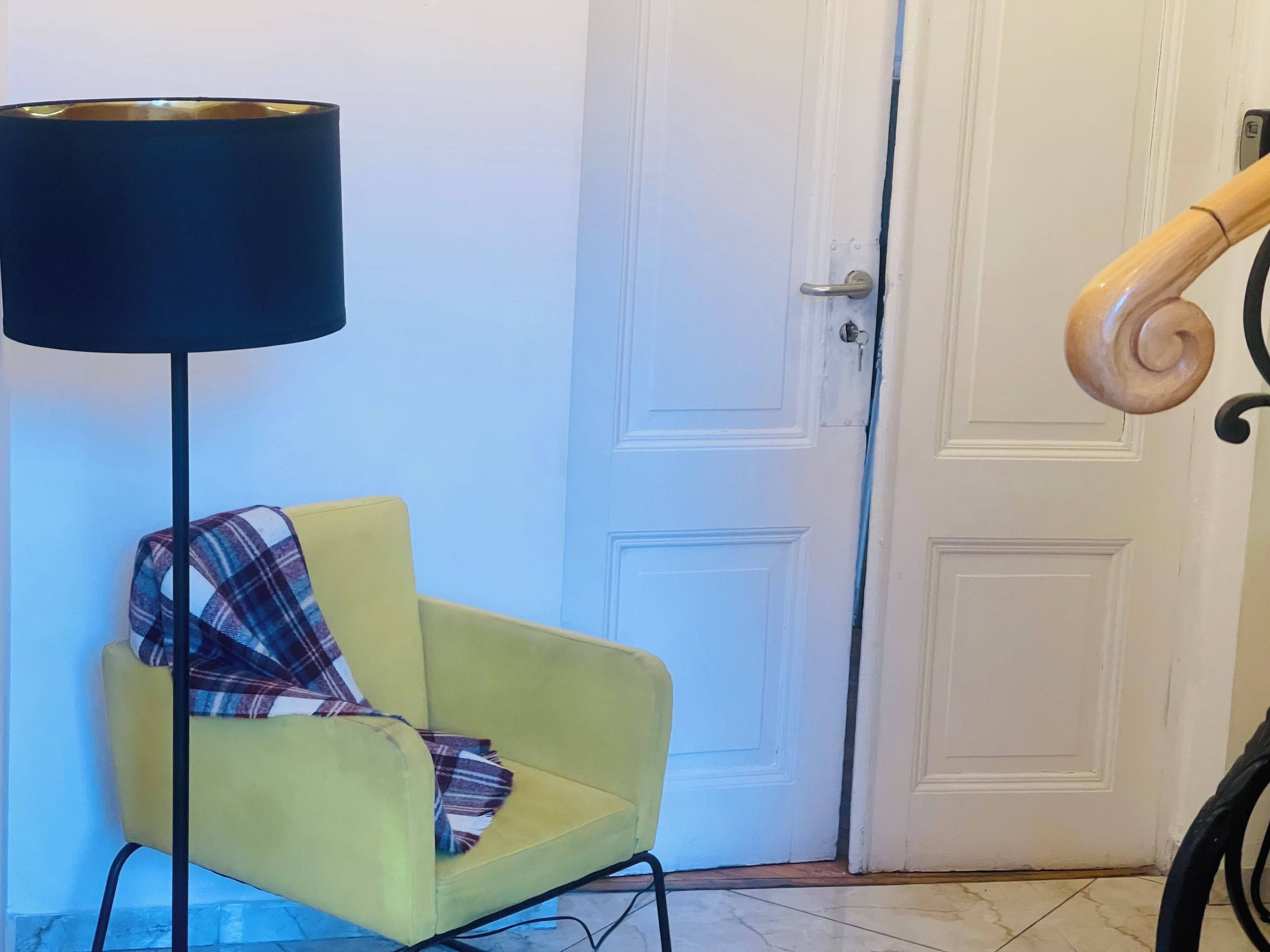 A yellow armchair with a plaid throw blanket, a floor lamp, and a white door with a wooden handrail.
