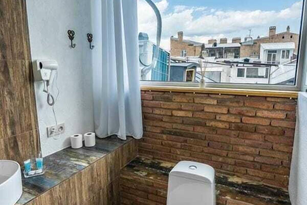 A modern bathroom with a toilet, a sink, and a shower area. It features a brick wall and a large window offering a view of buildings.