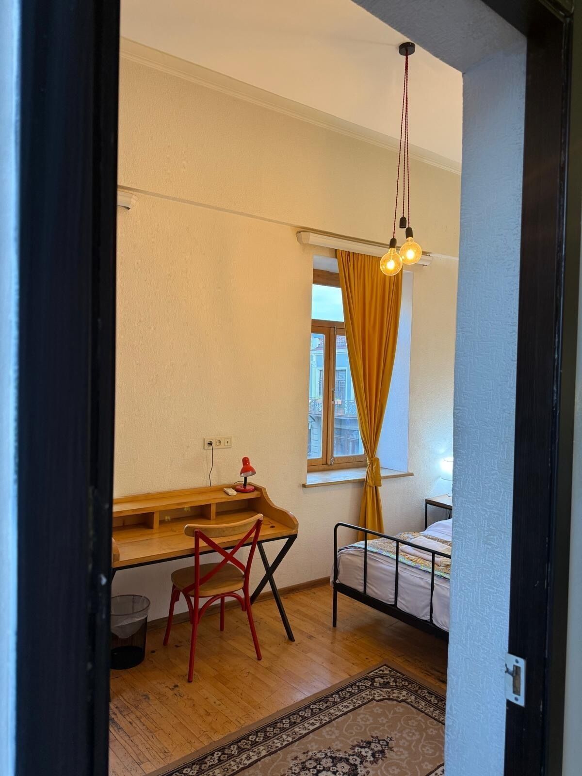 Quarto compacto com escrivaninha de madeira, uma cadeira vermelha, uma cama e um tapete estampado.