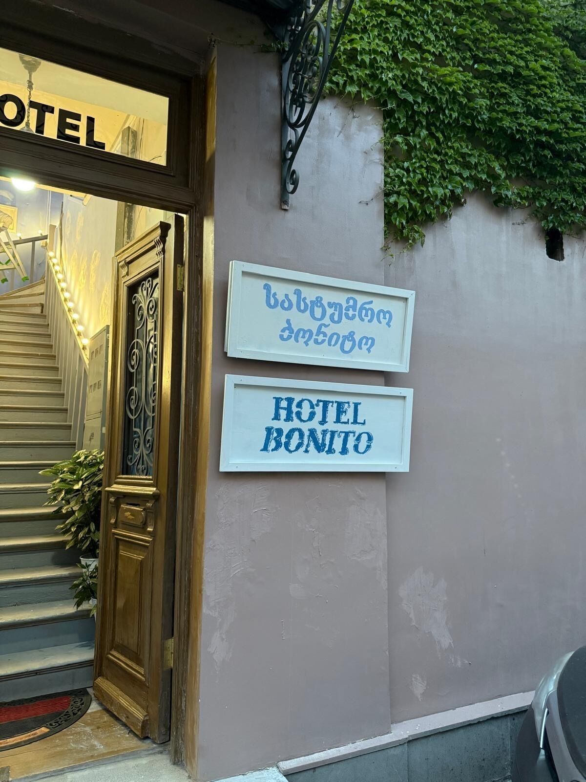 Entrada de hotel con una escalera, una puerta de madera y un letrero en la pared que indica el nombre del hotel tanto en inglés como en otro idioma.