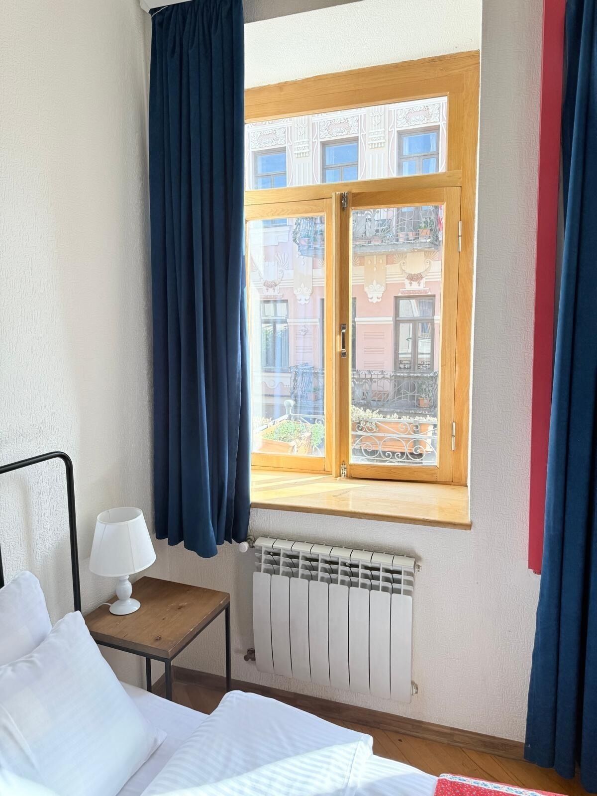 A bedroom with a bed, a bedside table with a lamp, a window with curtains, and a radiator.