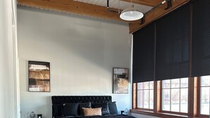 Een slaapkamer met een groot bed, houten balken in het plafond, twee nachtkastjes, een zwart hoofdeinde en een raam met jaloezieën.