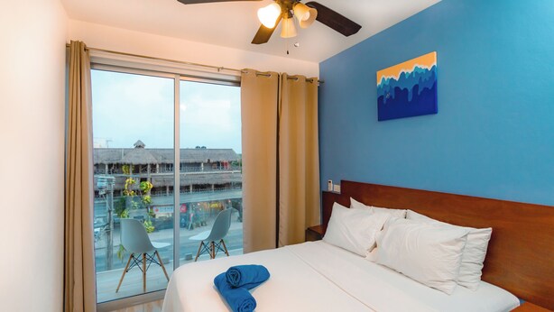 A hotel room with a large bed, a ceiling fan, a view of a building with a thatched roof, and a painting on the wall. - Charming 2-bedroom condo in Tulum with WiFi, AC (Tulum)