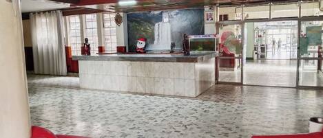 Um saguão de hotel com uma mesa de recepção, uma janela grande com cortinas e um piso ladrilhado.