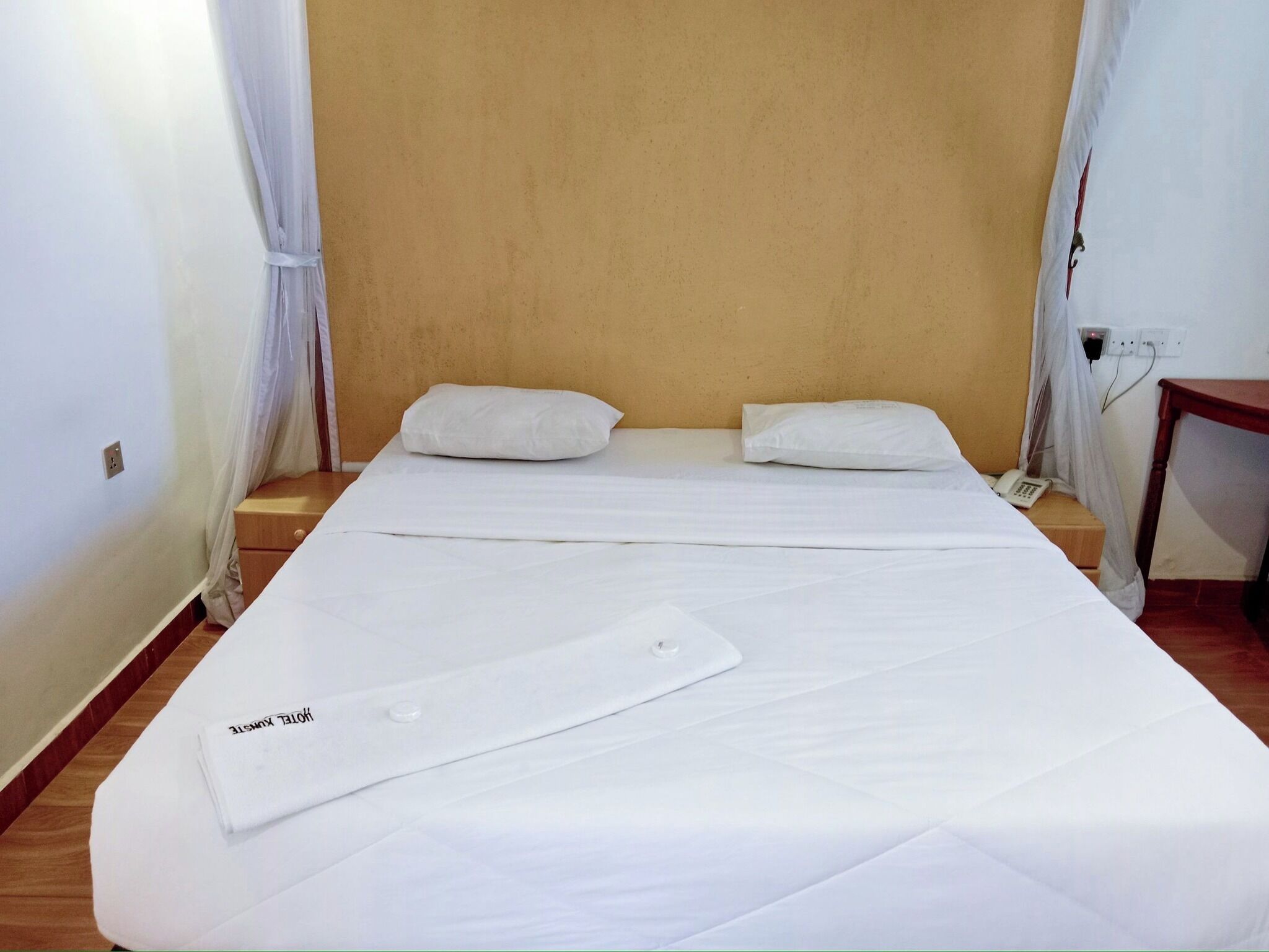 Habitación con una cama individual, una mesita de noche de madera, un teléfono y un enchufe en la pared.