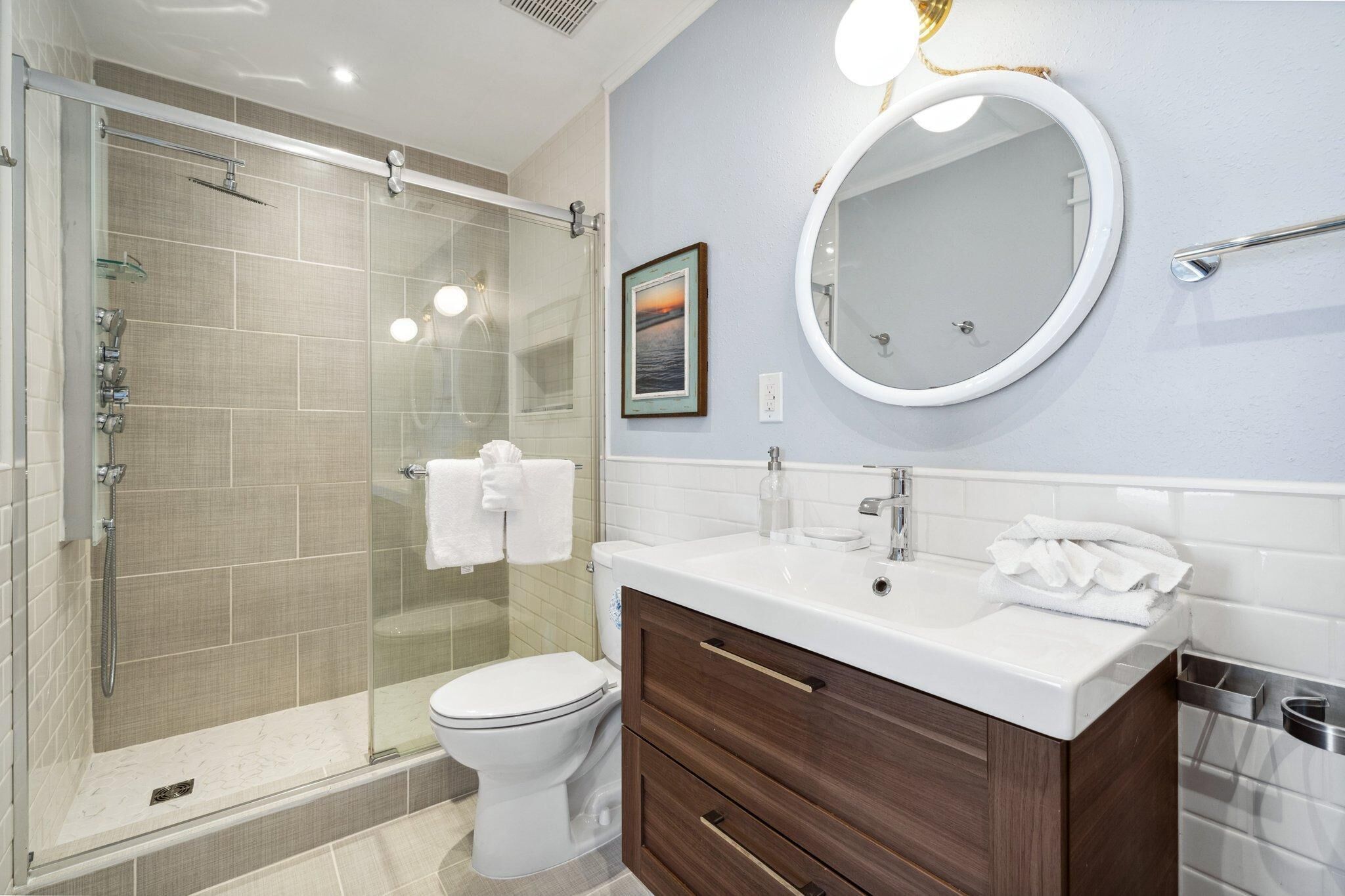 A modern bathroom with a glass-enclosed shower, a white toilet, a vanity with a mirror, and a framed picture on the wall.