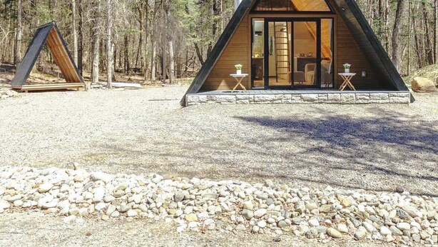 Een moderne, driehoekige houten cabin met een glazen deur, omgeven door een grindpad en dicht bos.