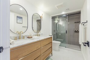 Un baño moderno con tocador de mármol, grifos dorados, cabina de ducha con mampara de vidrio y un inodoro blanco.