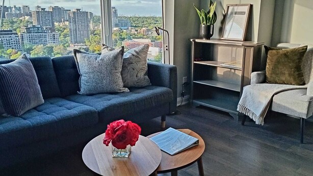 Un salon moderne avec un canapé bleu foncé, une table basse ronde ornée d’une rose rouge et une vue sur le paysage urbain, visible à travers de grandes fenêtres.