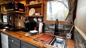 A compact kitchen with wooden cabinets, a built-in stove, and a window with curtains. - Custom HorseTrailerTinyHome near Grayson Highlands (Mouth of Wilson)