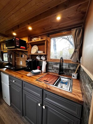 A compact kitchen with wooden cabinets, a built-in stove, and a window with curtains. - Custom HorseTrailerTinyHome near Grayson Highlands (Mouth of Wilson)
