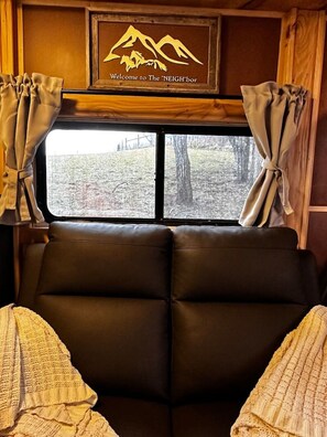 A cabin interior with a large window, curtains, and a sign reading "Welcome to The NEIGHBOR".