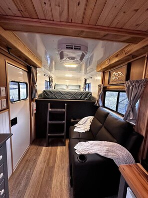 A compact RV interior with a kitchenette, a sleeping area with a sofa, and a small dining table.