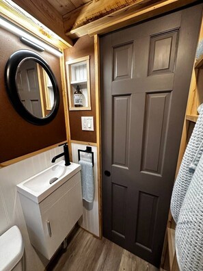 A bathroom with a white sink, black faucet, and a mirror. There is a towel hanging on a rack and a bottle of hand soap on the wall.