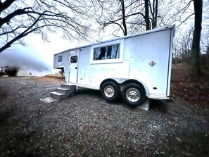 Une remorque blanche, avec une petite structure attenante, qui pourrait être une cuisine ou un espace de rangement, est garée sur une surface de gravier, avec des arbres en arrière-plan.