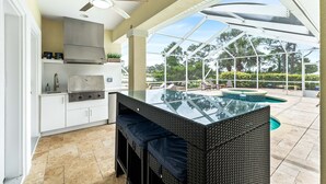 Une cuisine extérieure dotée d’un îlot central avec un plan de travail en verre, d’un ventilateur de plafond et offrant une vue sur une piscine et des arbres.