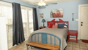 A bedroom with a bed, a bench, a ceiling fan, a window with curtains, a door, and a wall with a picture. - StoneFly Studio at Dragonfly Flats  Creede, Co (Creede)