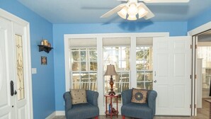 A living room with blue walls, two blue armchairs, a wooden coffee table, a ceiling fan, and a large window with a view of trees. - StoneFly Studio at Dragonfly Flats  Creede, Co (Creede)