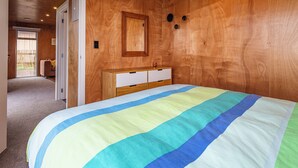 A bedroom with a wooden ceiling, a bed with a multicolored striped bedspread, a wooden dresser, and a door leading to another room. - Mountain Haus - Ohakune Holiday Home (Ohakune)