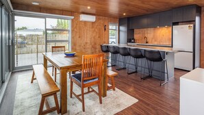 A modern kitchen with a wooden ceiling, a dining area with a wooden table and chairs, and a large window overlooking a backyard. - Mountain Haus - Ohakune Holiday Home (Ohakune)