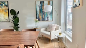 A modern living room with a wooden dining table, a white sofa, and a floor lamp. There are abstract paintings on the walls and a large window providing a view of the city.