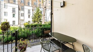 A balcony with a table and chairs, a potted plant, and a view of a building with multiple windows. - Stylish Bayswater Flat with Outdoor Space (London)