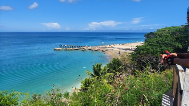 Une plage à l’eau d’un bleu limpide, avec une jetée et une végétation luxuriante.