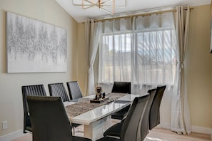 Uma área de jantar com mesa de vidro, cadeiras pretas, pintura abstrata branca, cortinas translúcidas e vista pela janela.