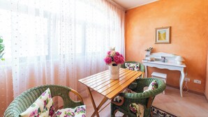 Una habitación con una mesa de madera, sillas de mimbre, un escritorio blanco y una ventana con cortinas traslúcidas.