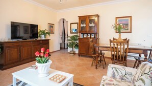 Una sala de estar con televisor, una mesa de centro blanca, un sofá floral, una mesa de comedor de madera y un aparador.