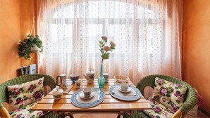 Un comedor con una mesa preparada para dos personas, con cojines de estampado floral, un jarrón con flores y una ventana con cortinas de encaje.