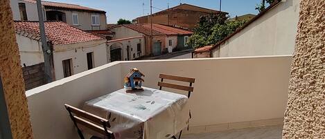 A small outdoor balcony with a table and chairs, a birdhouse, and a view of neighboring buildings.