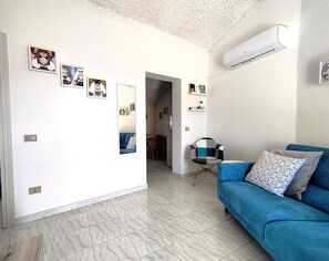 A living room with a blue sofa, a small table, and a chair. There are wall-mounted shelves with decorative items.
