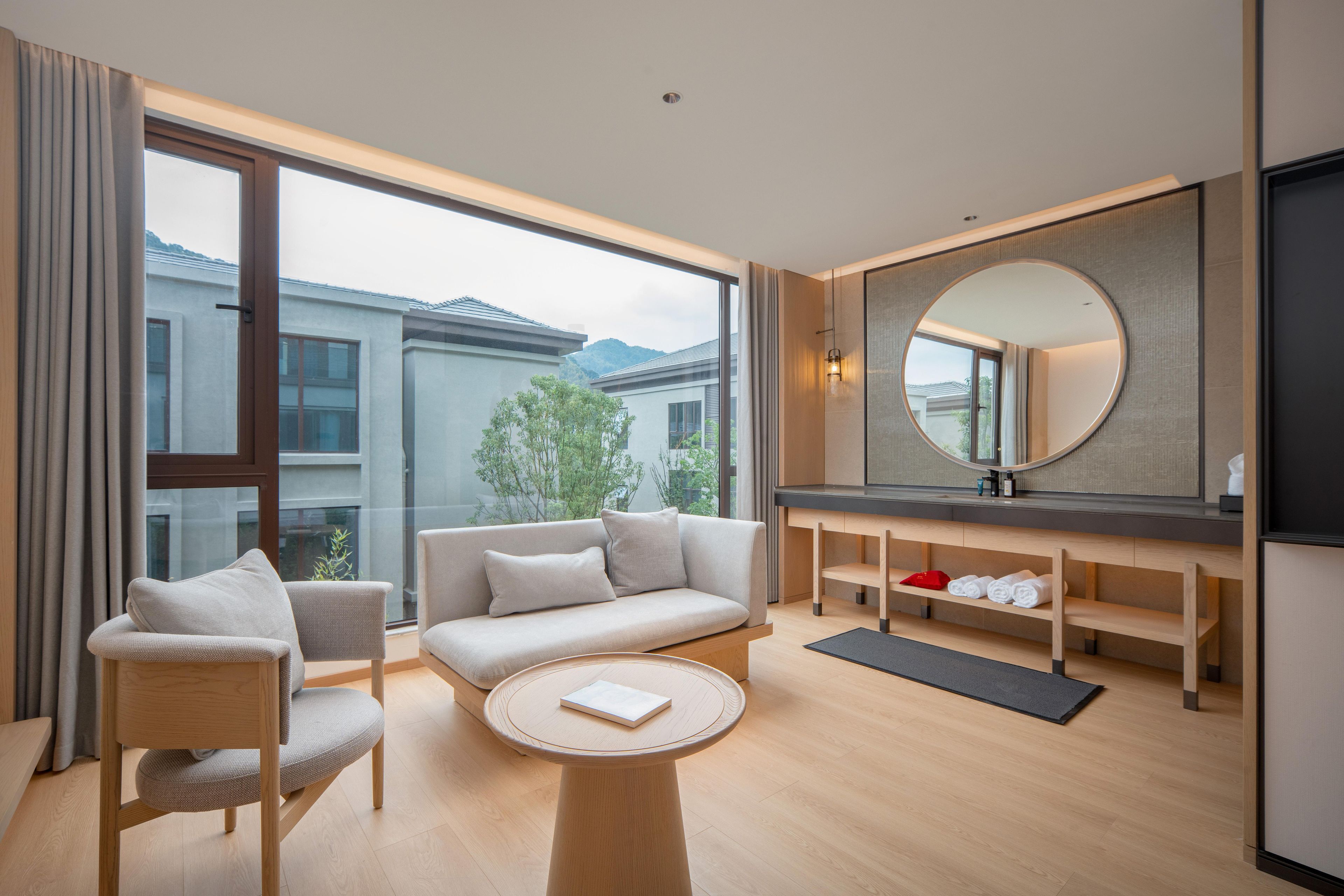 A modern hotel room with a sofa, chairs, a small table, a mirror, and a view of the outside.
