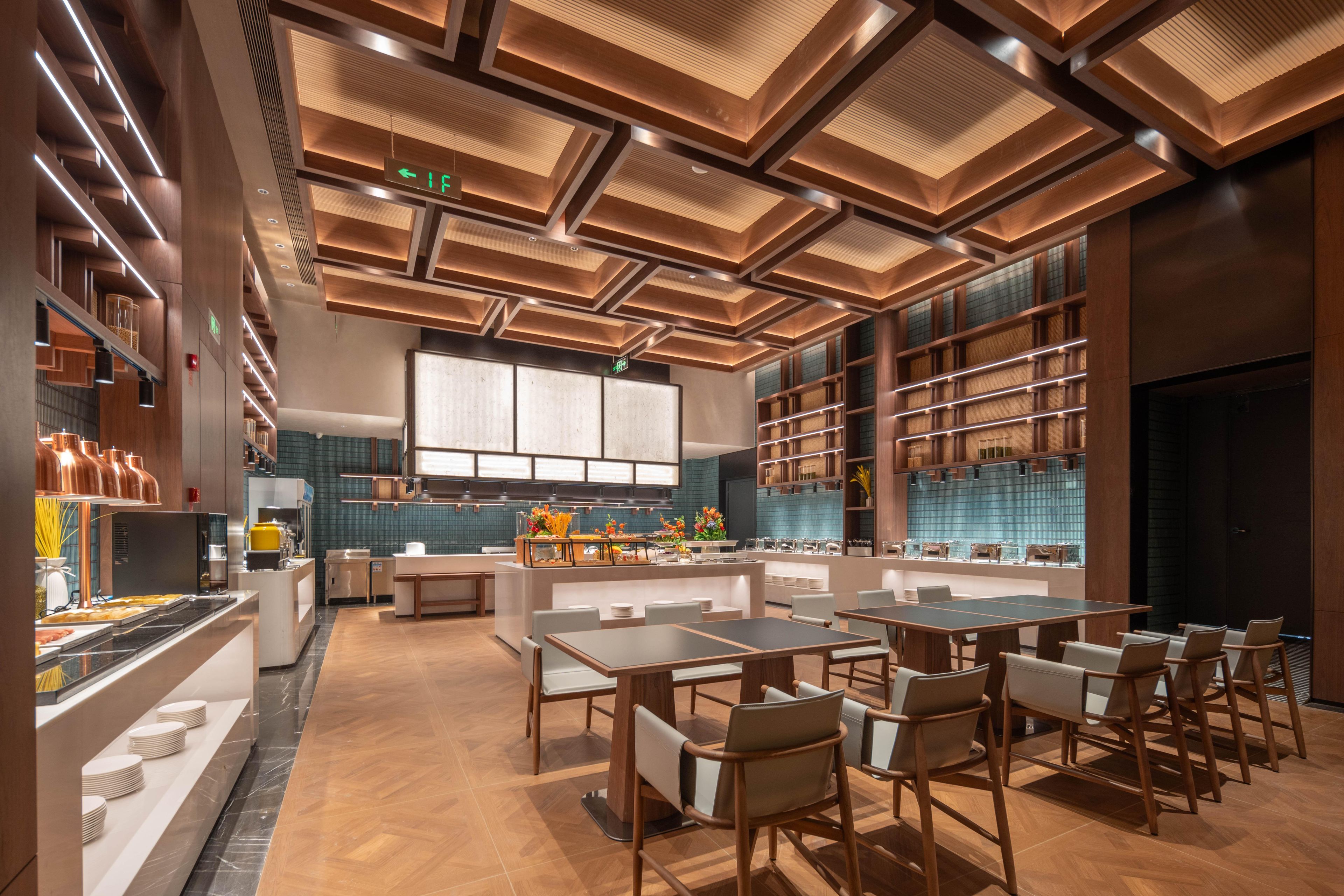 Een modern restaurant met houten vloeren, een centraal eetgedeelte met tafels en stoelen, een keuken met een groot raam en een plafond met inbouwverlichting.