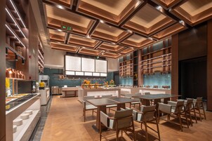 Un restaurante moderno con piso de madera, un área central de comedor con mesas y sillas, una zona de cocina con un ventanal grande, e iluminación empotrada en el techo.