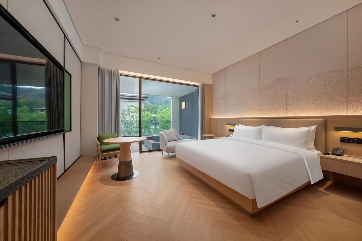A modern hotel room with a large bed, a small table, a chair, and a view of greenery through the window.