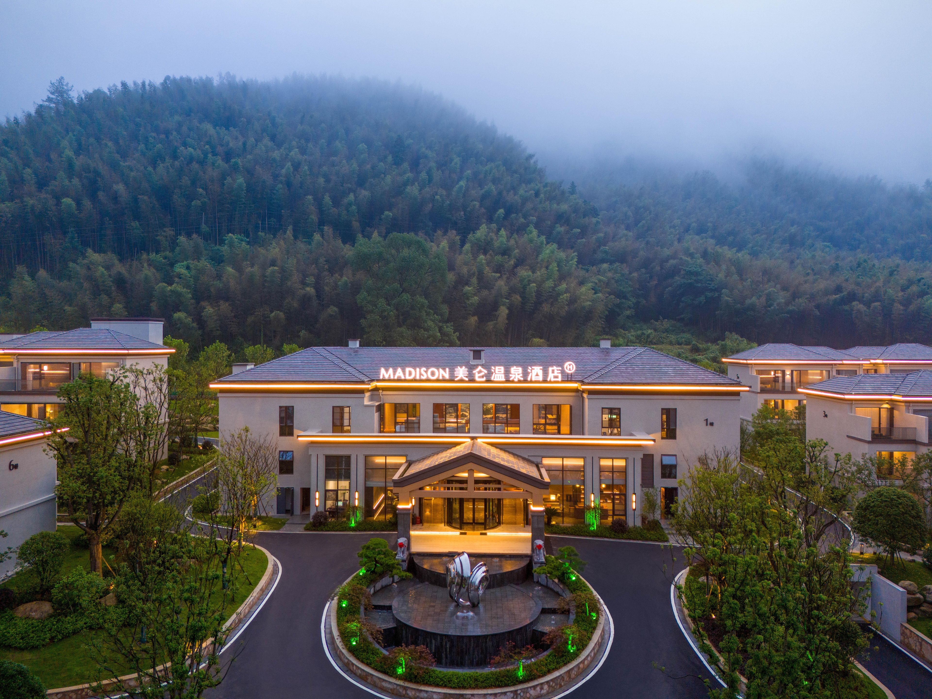 A hotel with a central building and smaller structures, surrounded by greenery and a mountainous backdrop.