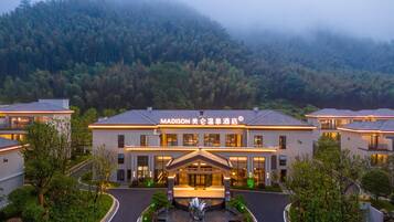 A hotel with a central building and smaller structures, surrounded by greenery and a mountainous backdrop.