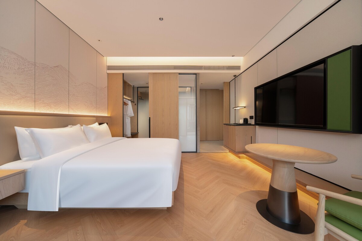 A modern hotel room with a large bed, a dining table, and a TV mounted on the wall.